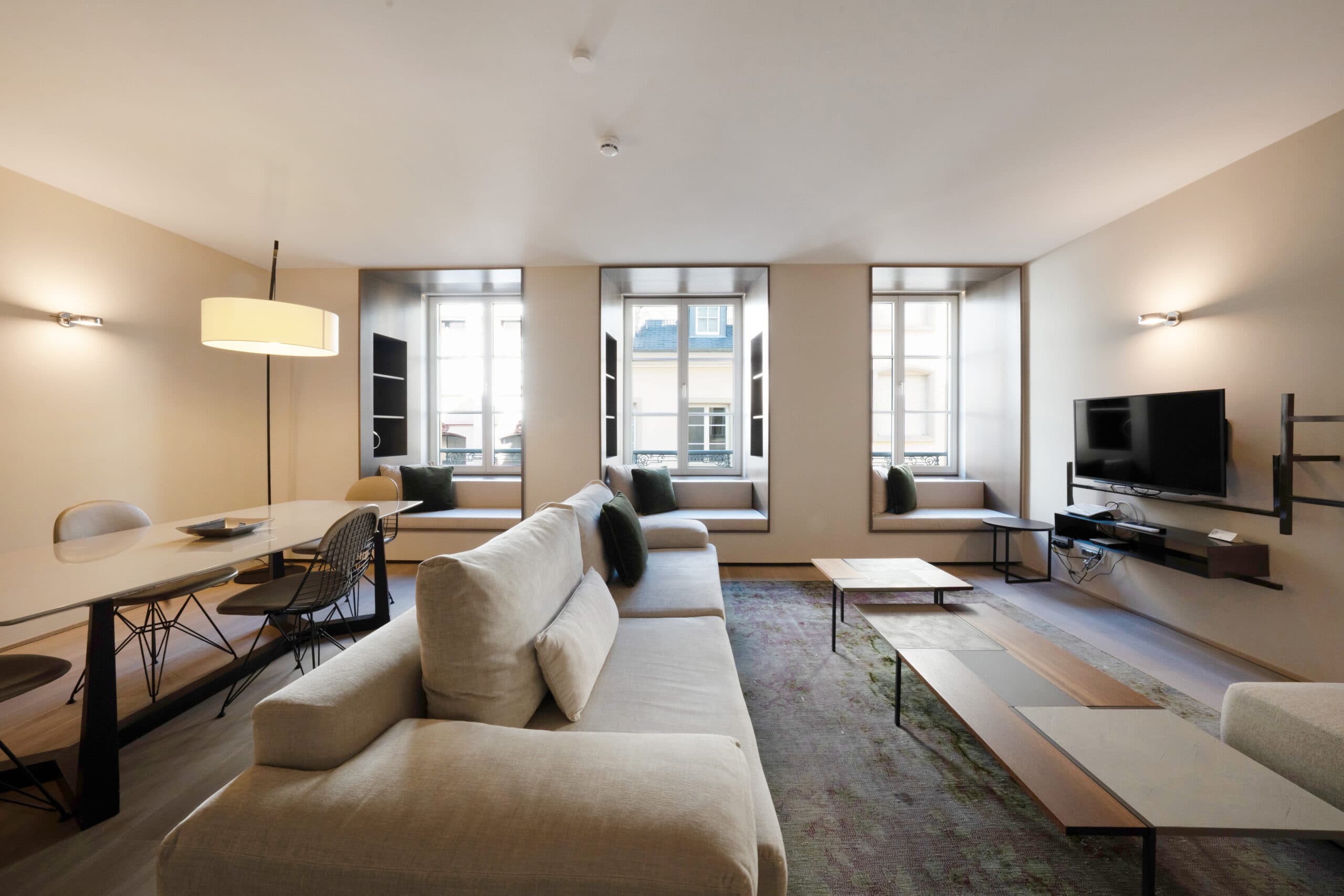 The wide living room of a one-bedroom apartment at Relais Louvigny, a sofa and dining table under tall windows.